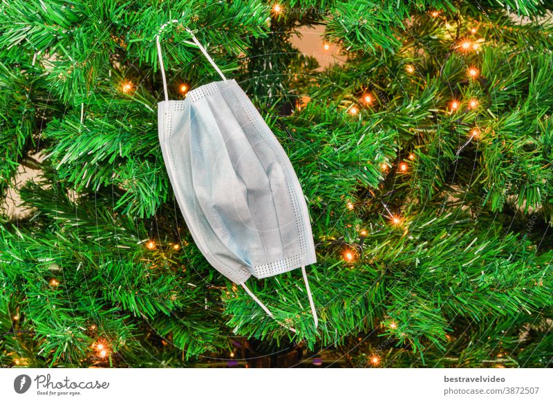 Weihnachtskuh-19 Maske, die als Dekoration an einem grünen Baum mit Lichtern hängt. Coronavirus-Gesichtsschutz schlichtes Design, das als jahreszeitlicher Schmuck an den Zweigen eines künstlichen Baumes verwendet wird.