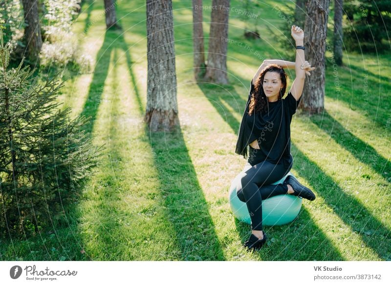 Foto einer aktiven, flexiblen, brünetten Frau, die sich mit Fitball streckt, Pilates oder Fitnessübungen macht, an einem sonnigen Frühlingstag im Wald auf grünem Gras posiert und an der frischen Luft gymnastische Übungen genießt.