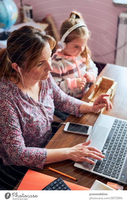 Mutter arbeitet bei der Erledigung ihrer Arbeit aus der Ferne während eines Video-Chat-Call-Stream-Online-Kurses Webinar auf einem Laptop von zu Hause aus, während ihre Tochter mit einem Ziegelsteinspielzeug spielt