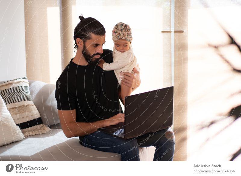 Vater mit Laptop, während er mit Baby auf dem Sofa sitzt multitask Arbeit heimwärts freiberuflich beschäftigt Kleinkind niedlich ethnisch Liege bezaubernd