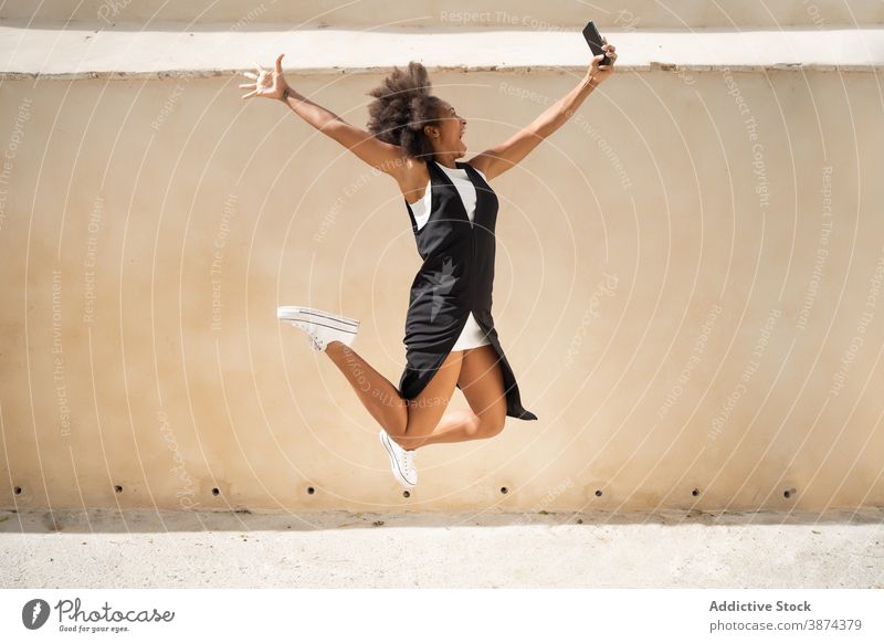 Positive ethnische Frau springt mit Smartphone auf die Straße springen sorgenfrei Selfie Spaß haben Freiheit ausdehnen aufgeregt Moment schwarz Afroamerikaner