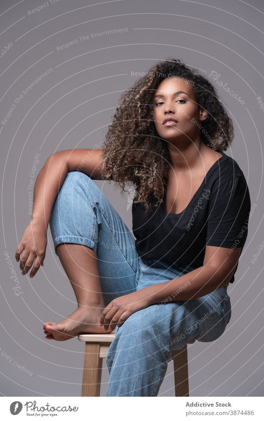 Stilvolle schwarze Frau in Freizeitkleidung sitzt auf Stuhl Mode Übergröße Übergewicht lässig Jeanshose Barfuß Model krause Haare jung Afroamerikaner ethnisch
