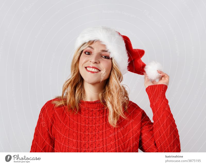 Lächelnde junge Frau in Santa Hut berührender Hut pom pom Weihnachtsmütze Hände Weihnachten Weihnachtsmann Bommel weiß vereinzelt rot grau abschließen Pullover