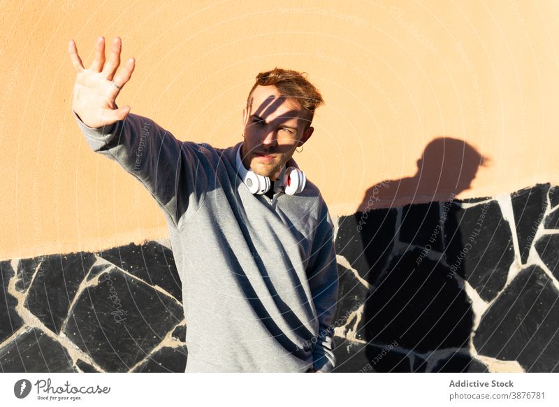 Junger Mann mit Kopfhörern, die das Gesicht vor Sonnenlicht schützen Sonnenschein Deckung behüten ausdehnen Hipster urban männlich modern unrasiert lässig Wand