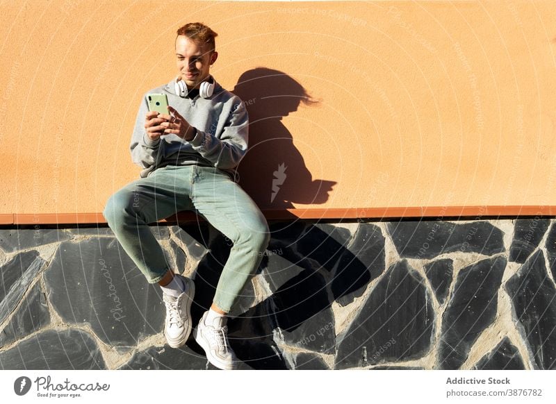 Junger Hipster-Mann beim Surfen auf dem Smartphone benutzend Wand urban Browsen positiv Telefon online jung Typ lässig Kopfhörer Mobile sonnig Schatten