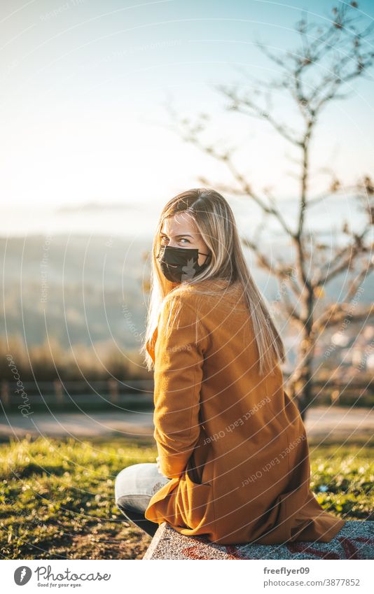 Frau im gelben Mantel auf einer Bank sitzend mit Gesichtsmaske Sitzen Coronavirus COVID Winter Herbst betrachtend Vigo Galicia Natur im Freien Freiheit