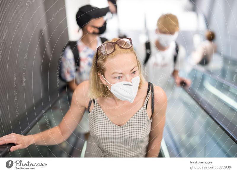 Porträt einer ungezwungenen jungen Frau, die im Kaufhaus Rolltreppen benutzt und eine Schutzmaske zum Schutz vor dem Covid-19-Virus trägt. Beiläufige Personen im Hintergrund