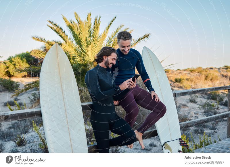 Fröhliche Surfer, die gemeinsam an der Böschung ihr Smartphone benutzen Männer benutzend Spazierweg Zusammensein Surfbrett Seeküste Stauanlage heiter Apparatur