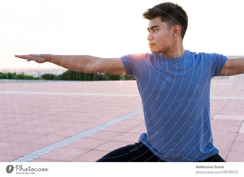 Ethnischer Mann in Sportkleidung macht Yoga bei Sonnenuntergang Krieger-Pose üben Asana beweglich Fokus ruhig männlich ethnisch asiatisch virabhadrasana urban
