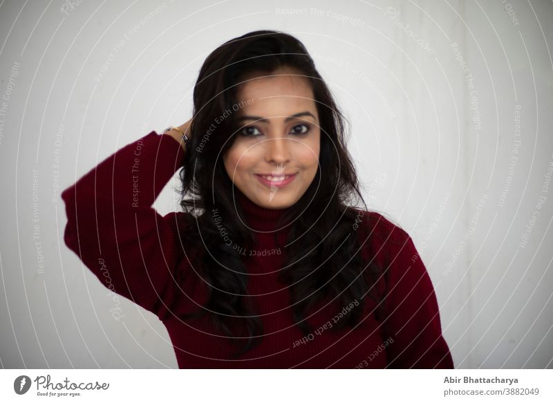 Porträt einer jungen, lächelnden, brünetten, indischen Bengalinerin in kastanienbraunem, westlichem Wollpullover mit hohem Halsausschnitt, der in einem weißen Copy-Space-Hintergrund an die Wand gelehnt ist. Indischer Lebensstil und Winterkollektion