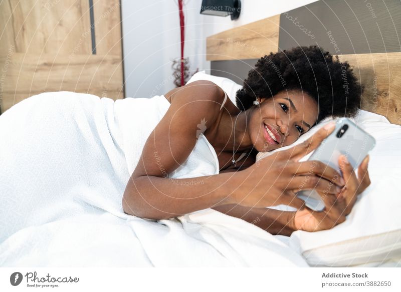 Schwarze Frau benutzt Smartphone im Bett Browsen Morgen wach benutzend Lächeln Süchtige soziale Netzwerke ethnisch schwarz Afroamerikaner Telefon Mobile