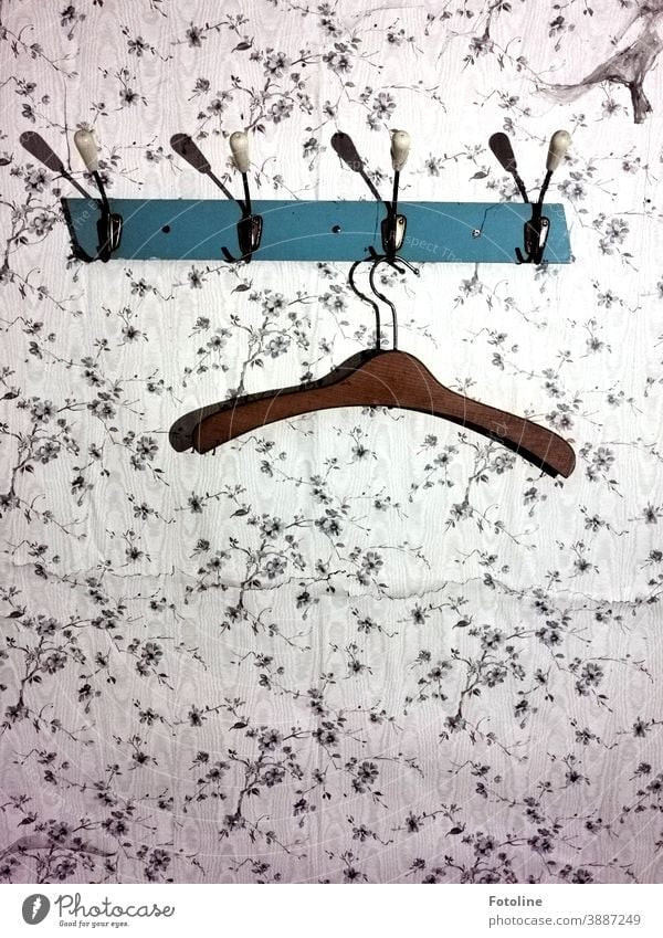 Ein Bügel hängt an einer alten Garderobe die auf einer gemusterten Tapete angebracht ist. Garderobenhaken Kleiderbügel Haken lost places Muster Riss Holz Metall