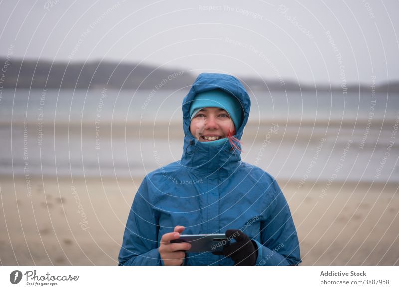 Fröhlich reisende Frau mit Smartphone in der Nähe von Meer im Herbst Reisender MEER Entdecker warme Kleidung Wind Meeresufer heiter wasserdicht Jacke