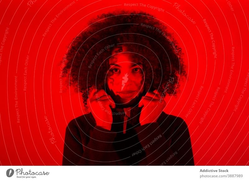 Lockig behaarte ethnische Frau in roter Neon-Beleuchtung krause Haare neonfarbig jung leuchten Licht Farbe Porträt trendy Afroamerikaner schwarz Vorschein