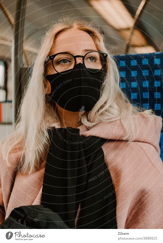 Junge Frau sitzt mit Coronamaske in der Bahn coronavirus junge frau blond lange haare bahn reise gesellschaft winter mantel brille mode style blick hübsch schön