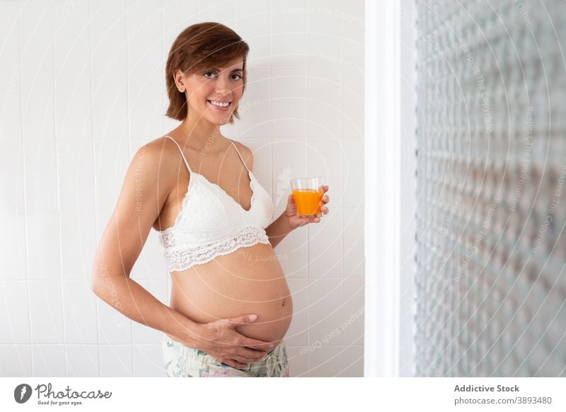 Lächelnde schwangere Frau mit Glas Saft zu Hause Gesundheit trinken Schwangerschaft heimwärts frisch orange BH erwarten Bauch mütterlich charmant vorwegnehmen