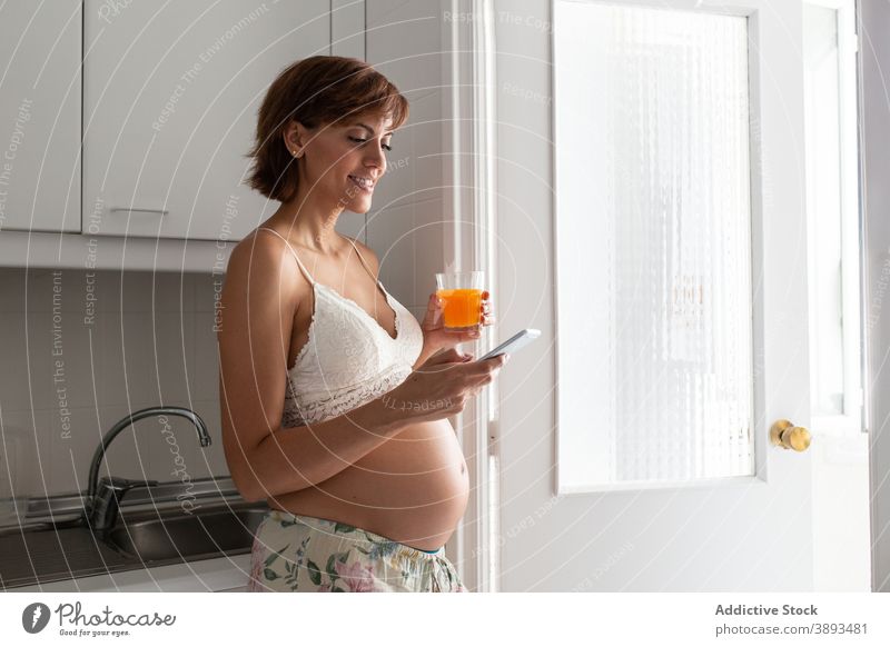 Zarte schwangere Frau mit Smartphone in der Küche Browsen heimwärts Schwangerschaft Angebot Nachricht Lächeln Apparatur Gerät pränatal Surfen Mutterschaft