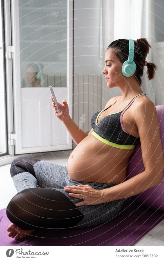 Schwangere Frau in Sportkleidung am Mobiltelefon zu Hause schwanger Yoga Schwangerschaft Smartphone Unterlage Bauch zuhören Musik benutzend Kopfhörer Apparatur