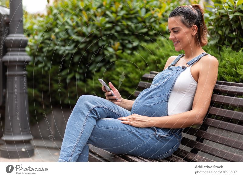Lächelnde schwangere Frau mit Smartphone in der Stadt Großstadt Browsen Straße Bank hölzern Apparatur Windstille Surfen Anschluss online Kommunizieren Unterleib