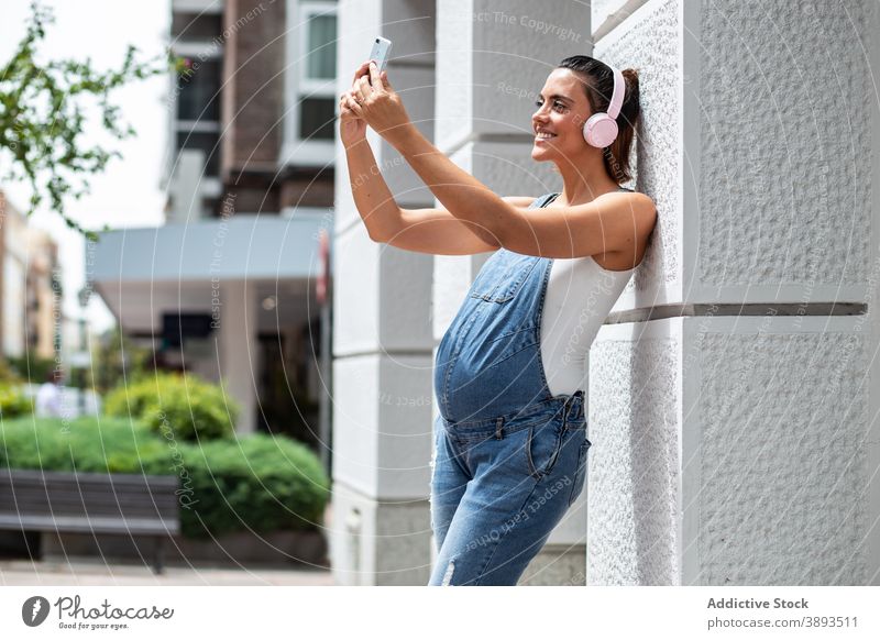 Freudige schwangere Frau nimmt Selfie in der Stadt Schwangerschaft Großstadt schlendern Kopfhörer Musik Selbstportrait heiter Straße urban Gebäude Foto Mobile