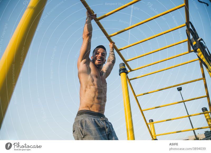 Athletischer junger Mann, der in der Gymnastikhalle im Freien an den Stangen hängt calisthenics Sport Muskeln Stärke erhängen Glück lustig Lächeln Kunstturnen