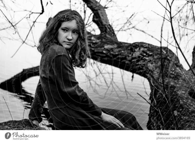 Melancholisch und etwas vorwurfsvoll blickt das Mädchen am Seeufer sich um junge Frau Teenager nachdenklich ernst Ufer Baum Baumstamm Kleid dunkel düster