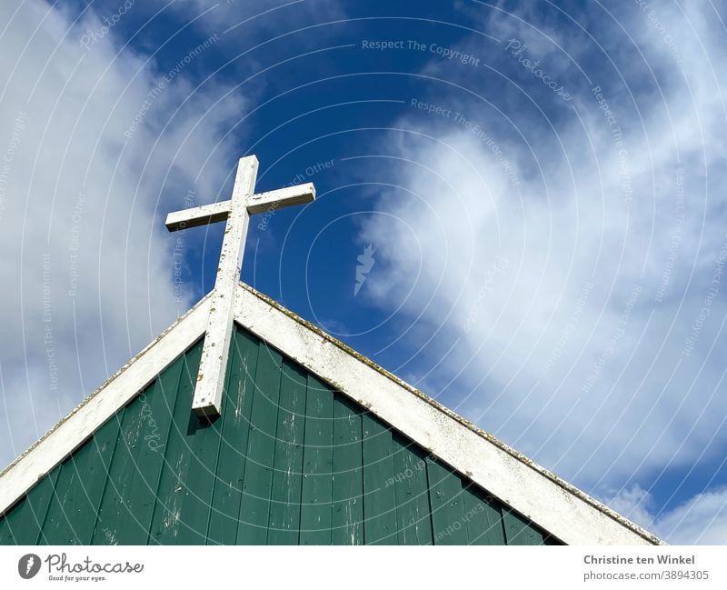 Weißes Kreuz aus Holz an der Giebelwand einer Kapelle, die mit grünem und weissem Holz verkleidet ist.  Alles ist etwas verwittert. Blick von unten gegen den blauen bewölkten Himmel. Spiekeroog I Farbkombination