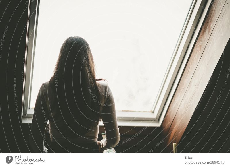 Rückansicht einer Frau mit langen dunklen Haaren die aus einem Fenster in einer Dachschräge blickt Dachfenster rausschauen Rücken lange Haare von hinten zuhause