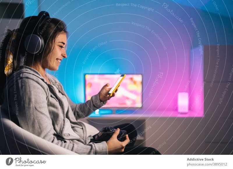 Lächelnde weibliche Gamerin verwendet Smartphone zu Hause Spieler Frau Gamepad Regler Konsole Browsen Joystick Videospiel heiter Abend heimwärts Kopfhörer