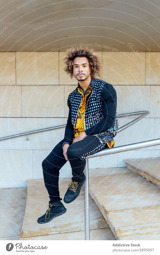 Stilvolle Hipster ethnischen Mann sitzt auf Geländer trendy informell Mode krause Haare Vollbart jung modern Outfit Schritt Reling Typ Afroamerikaner schwarz