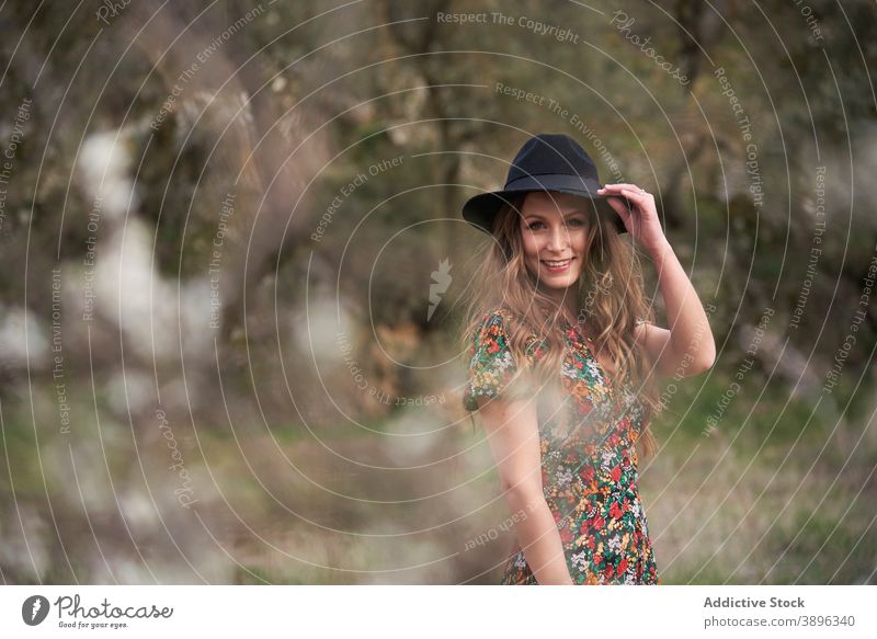 Fröhliche Frau mit Hut im Wald stehend Accessoire charmant Kleid Lächeln Outfit Natur Bekleidung Wälder Vorschein lange Haare gewelltes Haar Optimist Stil