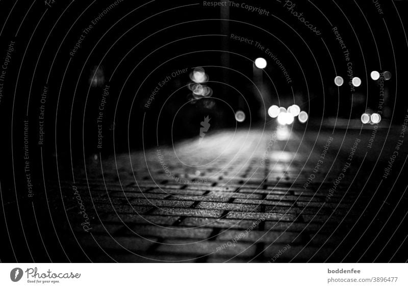 reflektierendes Licht einer Straßenlaterne auf dem gepflasterten Gehweg, im Hintergrund ein Bokeh von Autolichtern und Straßenlaternen, fokussiert auf den Vordergrund, Froschperspektive