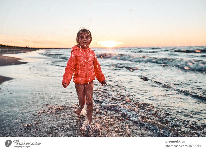 Verspieltes kleines Mädchen, das mit Wasser in Richtung Kamera spritzt und bei Sonnenuntergang an einem Sandstrand über dem Meer seine Freizeit genießt