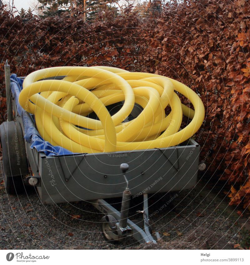 gelbe Plastik-Leerrohre liegen zusammengerollt auf einem Anhänger, der vor einer Buchenhecke steht Kabelschutzrohr flexibel Tiefbau lang Schutzrohr Hecke Plane