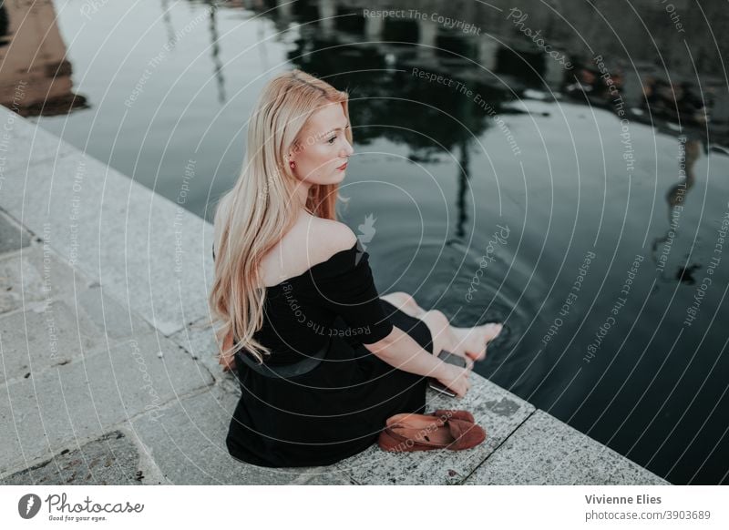 Frau mit nackten Füßen am Wasser sitzend blond lange Haare elegant schwarzes Kleid Meditation entspannend Natur See Fluss Baden baden Denken nachdenklich