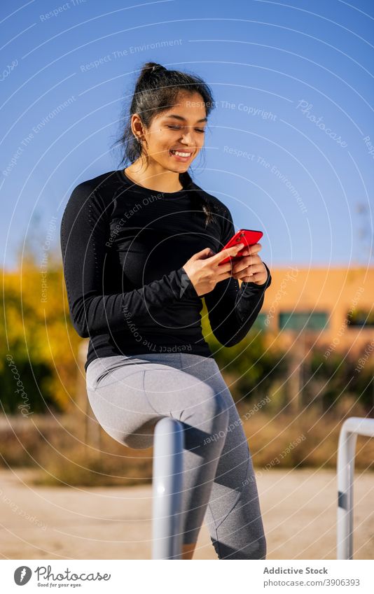 Lächelnde ethnische Sportlerin beim Surfen im Smartphone auf dem Sportplatz Browsen Pause Training calisthenics parallel Bar sich[Akk] entspannen Frau