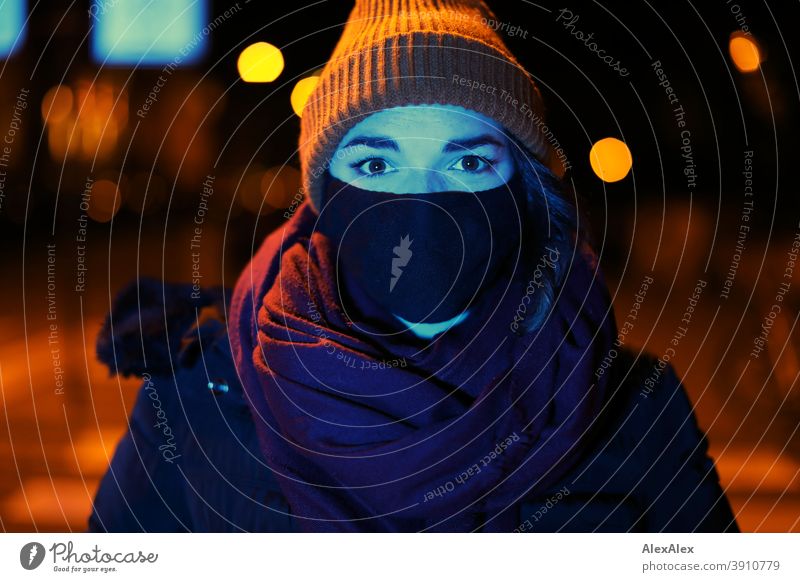 Junge Frau mit Maske und Mütze steht nachts mit blauem Licht angeleuchtet in orangem Laternenlicht in der Stadt Dschungel Jugendliche blaues Licht Straße