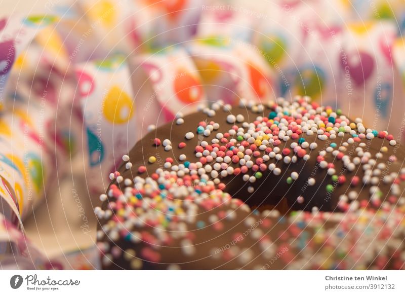 Donut / Doughnut mit Schokolade und Zuckerperlen. Dahinter Luftschlangen mit Luftballon Motiv in Nahaufnahme. Schwache Tiefenschärfe mit Fokus auf den Zuckerperlen