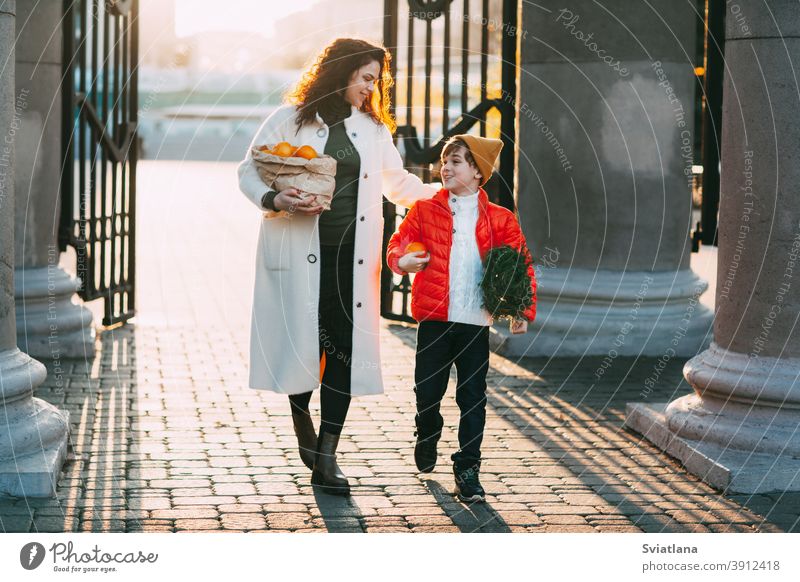 Eine schöne Mutter mit ihrem Sohn in einer leuchtend orangefarbenen Jacke und einem modischen gelben Hut gehen aus dem Laden und halten ein Paket mit Orangen und einem kleinen Weihnachtsbaum. Einkaufen für die Feiertage. Vorbereitungen für Weihnachten und Neujahr