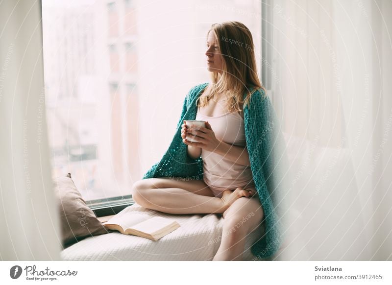 Ein junges Mädchen sitzt frühmorgens auf der Fensterbank, trinkt heißen Kaffee, und neben ihr liegt ein aufgeschlagenes Buch Frau Fensterbrett Sitzen Freizeit