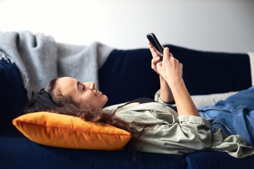 Junge lächelnde Frau entspannt sich zu Hause auf dem Sofa liegend und überprüft soziale Medien auf dem Handy Lügen räkeln Mobile Zelle Smartphone Technologie