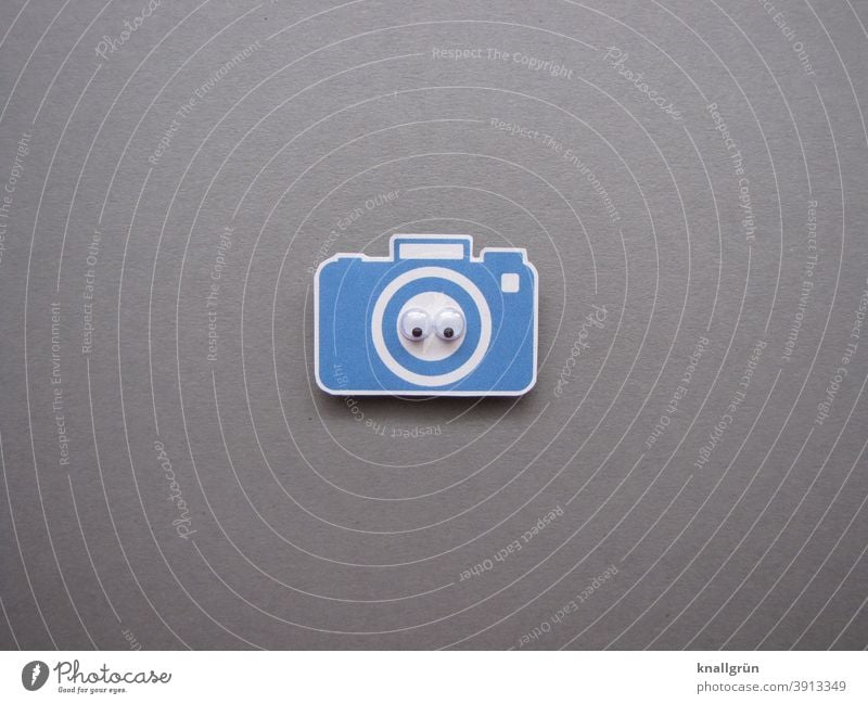 Kleine blaue Kamera aus Papier mit zwei Wackelaugen Blick Auge Blick in die Kamera ausgeschnitten Farbfoto weiß grau schwarz Zentralperspektive Nahaufnahme
