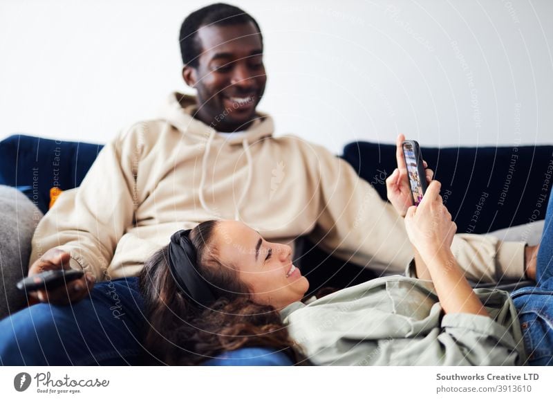 Entspanntes junges Paar zu Hause auf dem Sofa sitzend, fernsehend und soziale Medien auf dem Mobiltelefon überprüfend Sitzen räkeln zuschauend FERNSEHER