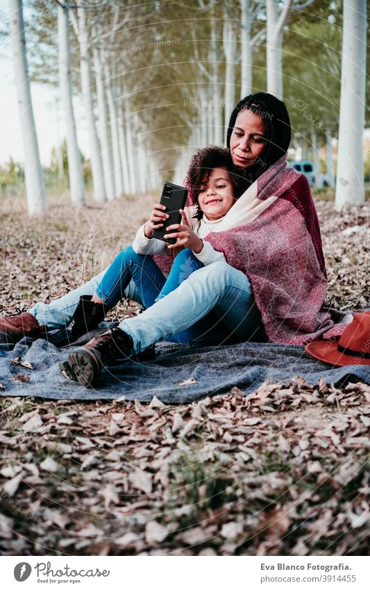 hispanische Mutter und afro Kind Mädchen im Freien umarmen bei Sonnenuntergang in Decke eingewickelt. Herbstzeit. Familie und Liebe Konzept Handy