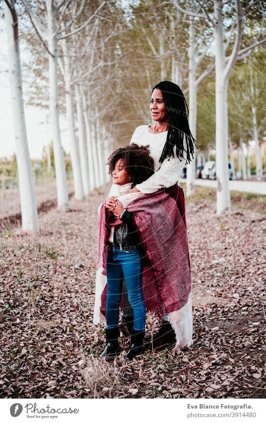 hispanische Mutter und afro Kind Mädchen im Freien umarmen bei Sonnenuntergang in Decke eingewickelt. Herbstzeit. Familie und Liebe Konzept Umarmung Afro-Look