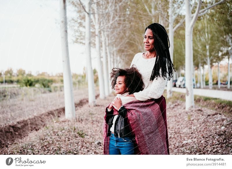 hispanische Mutter und afro Kind Mädchen im Freien umarmen bei Sonnenuntergang in Decke eingewickelt. Herbstzeit. Familie und Liebe Konzept Umarmung Afro-Look