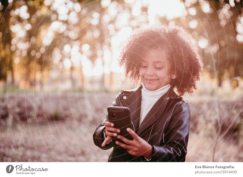 Porträt von niedlichen Afro Kind Mädchen mit Handy bei Sonnenuntergang während der goldenen Stunde, Herbstzeit, schöne Bäume Hintergrund Technik & Technologie