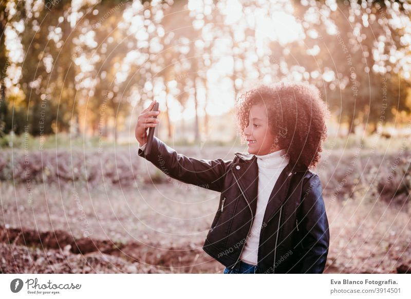 Porträt von niedlichen Afro Kind Mädchen mit Handy bei Sonnenuntergang während der goldenen Stunde, Herbstzeit, schöne Bäume Hintergrund Technik & Technologie