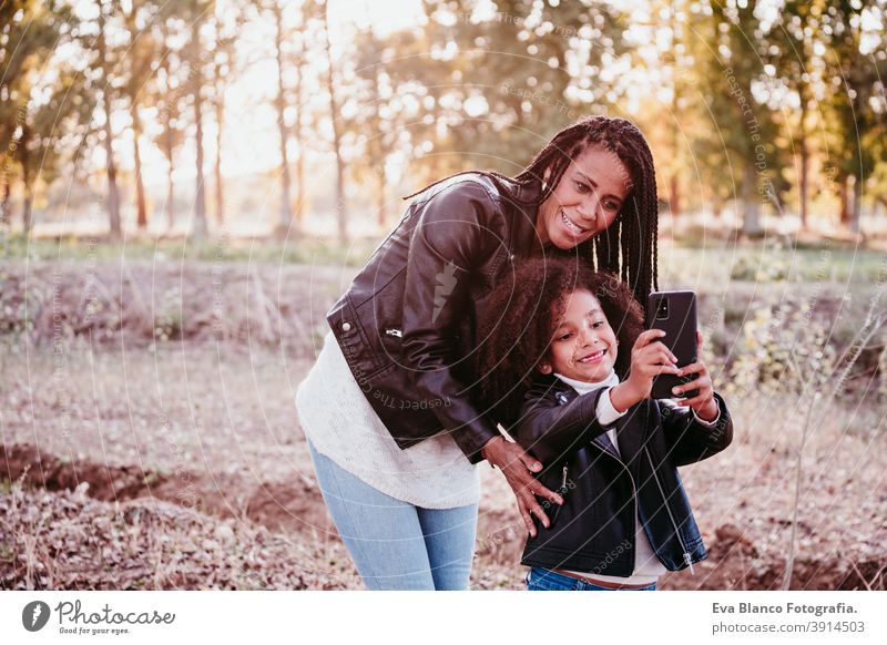 hispanische Mutter und niedliche afro Kind Mädchen nehmen selfie im Freien bei Sonnenuntergang. Familie, Liebe und Technologie-Konzept Handy Selfie Gerät