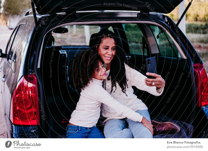 hispanische Mutter und afro Kind Mädchen sitzen in einem Auto in der Natur. Sie haben Spaß und machen Fotos mit dem Mobiltelefon. Herbst-Saison. Familie und Reisen Konzept